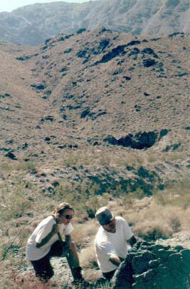 photo of Jim banging on an outcrop with his hammer as Paula looks on; desert hills in background